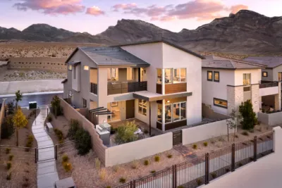 Modern two-story house with large windows and balconies, surrounded by a low fence, desert landscaping, and sidewalks; mountains and a dramatic sky are visible in the background.