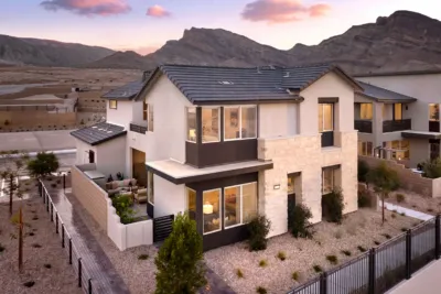 Modern two-story house with large windows, stone and stucco exterior, and a fenced yard, set in a desert landscape with mountains in the background at sunset.