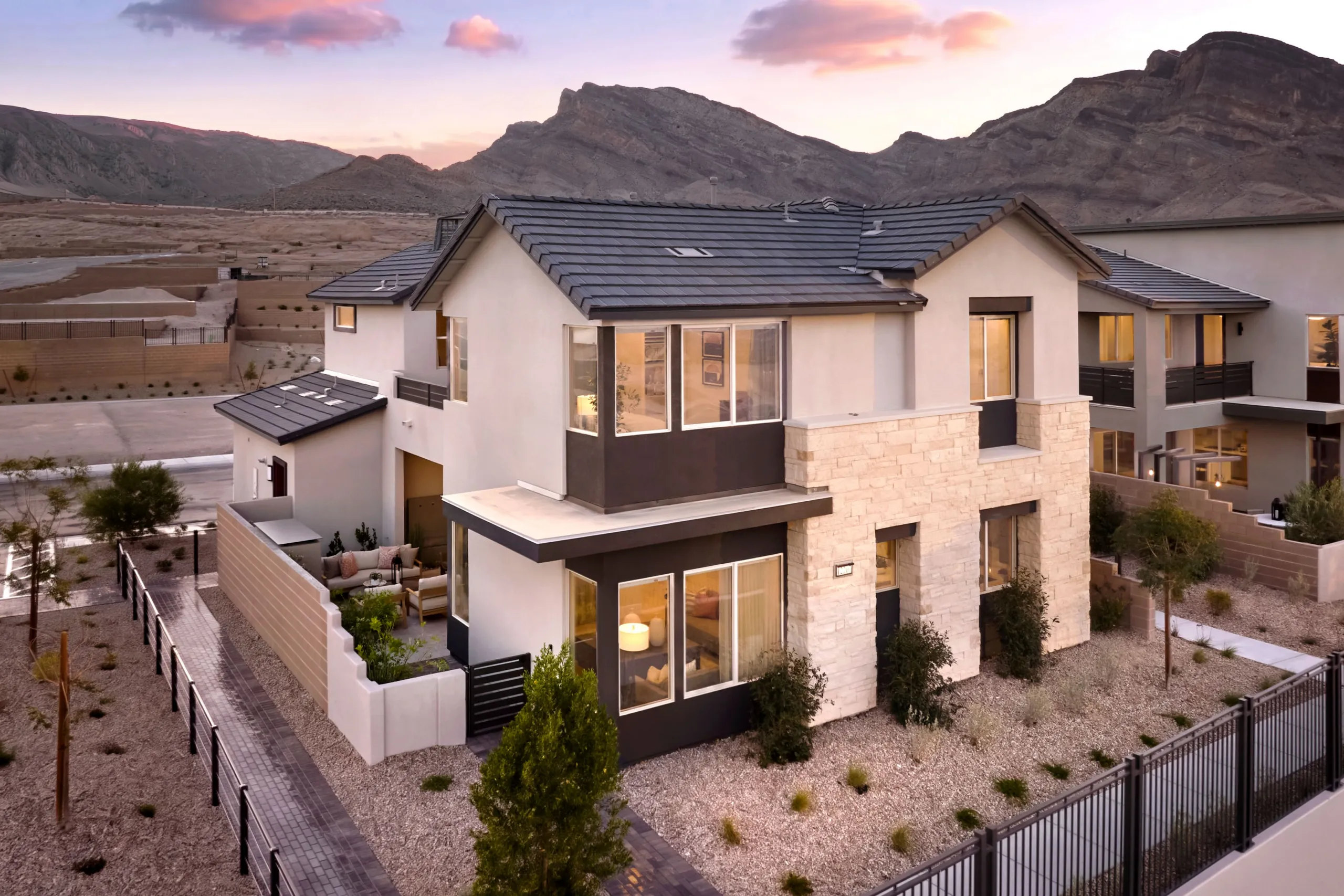 Modern two-story house with large windows, stone and stucco exterior, and a fenced yard, set in a desert landscape with mountains in the background at sunset.