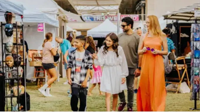 A group of four people, including two children and two adults, walk and smile together at an outdoor market with art displays, stalls, and various shoppers in the background.