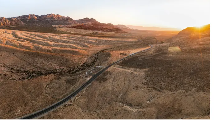 Aerial view of a long, winding road cutting through a vast, arid desert landscape with rocky hills in the distance and the sun setting on the horizon, casting a warm golden light.