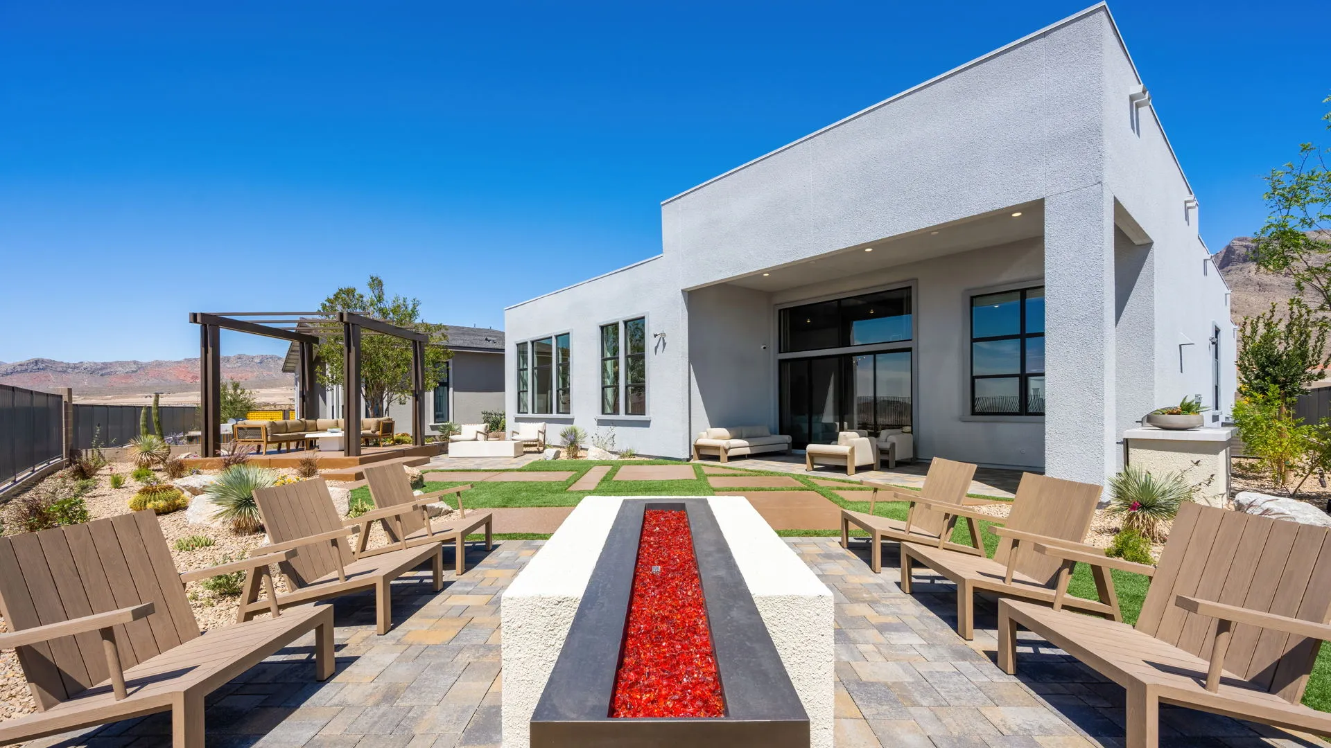 Backyard of Monroe Model at Esplanade at Red Rock by Taylor Morrison (4) Modern backyard patio with wooden chairs surrounding a long rectangular fire pit with red glass, desert landscaping, and a white house under a clear blue sky.