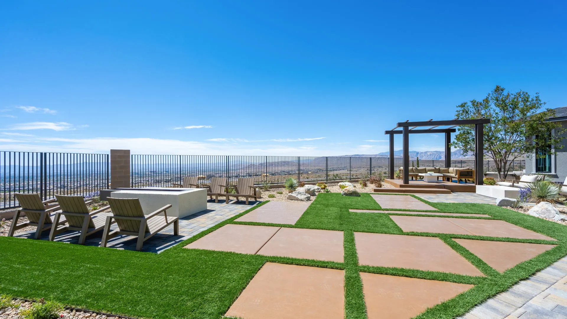 Backyard of Monroe Model at Esplanade at Red Rock by Taylor Morrison A modern backyard features large pavers in artificial grass, patio seating under a pergola, lounge chairs by a low fence, and mountain views under a clear blue sky.
