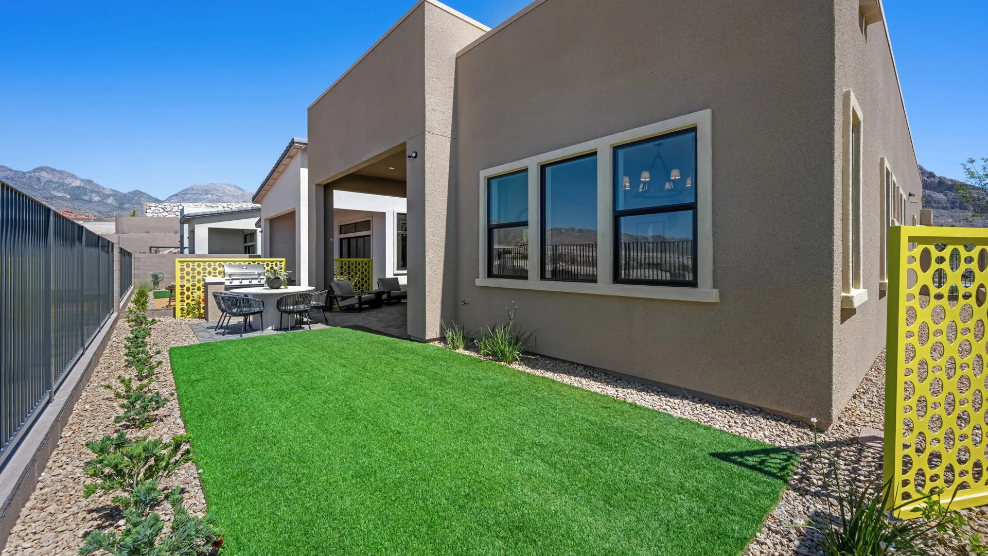 Backyard of Paloma Model at Esplanade at Red Rock by Taylor Morrison (1) Modern backyard with artificial grass, gravel, and shrubs beside a beige house. There is an outdoor dining area under a covered patio, decorative yellow panels, and mountains visible in the background under a clear blue sky.