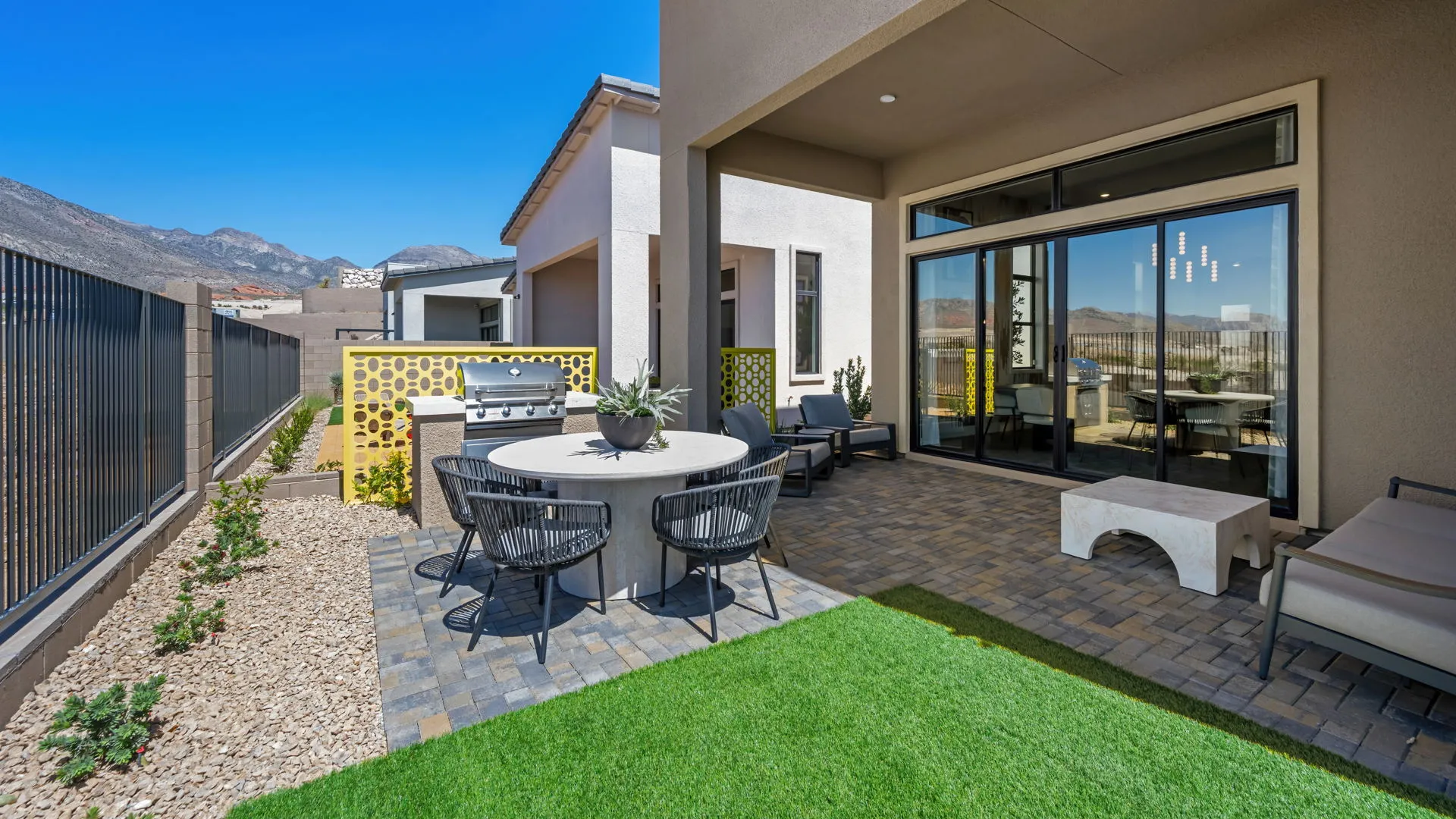 Backyard of Paloma Model at Esplanade at Red Rock by Taylor Morrison (2) Modern backyard patio with a round dining table, four black chairs, potted plants, grill, and outdoor seating area. Green artificial turf and brick pavers cover the ground; mountains visible in the background under a clear blue sky.