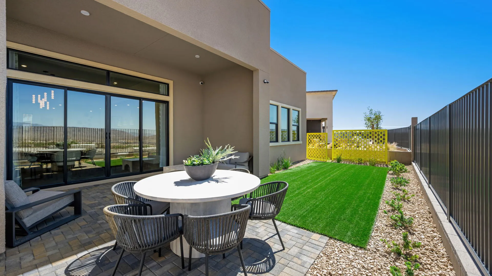 Backyard of Paloma Model at Esplanade at Red Rock by Taylor Morrison Modern backyard patio with a round dining table and chairs, artificial grass, stone landscaping, and a yellow decorative privacy screen against a blue sky.