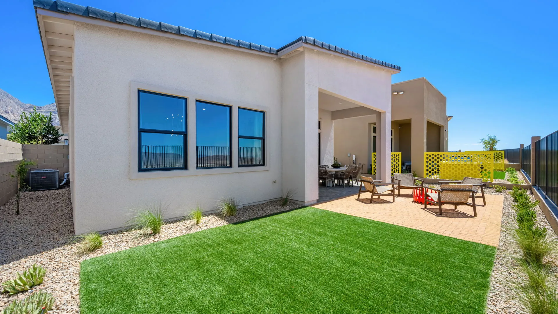 Backyard of Sangria Model at Esplanade at Red Rock by Taylor Morrison Modern house with large windows, a covered patio with dining area, and a backyard featuring artificial grass, a paved seating area, desert landscaping, and yellow privacy screens under a clear blue sky.