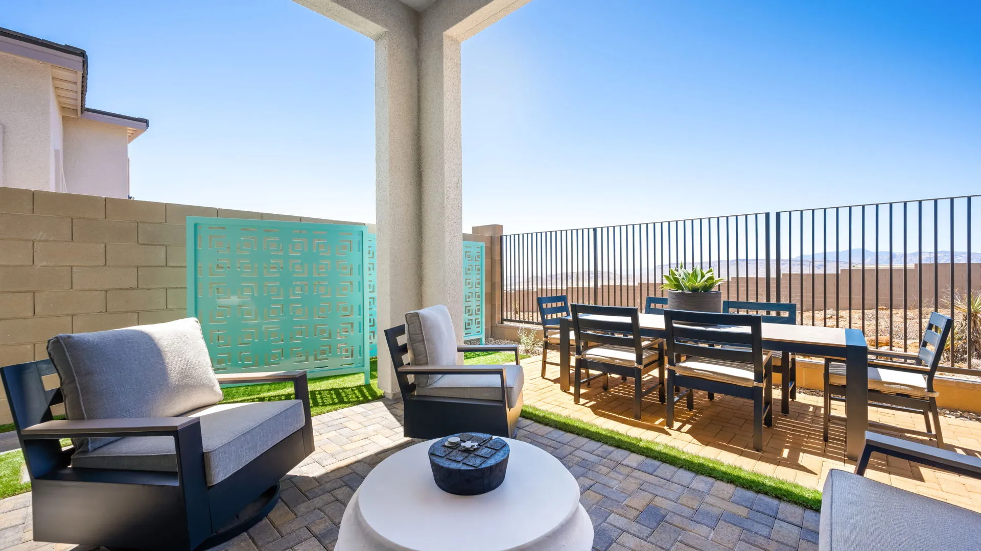 Backyard of Tiki Model at Esplanade at Red Rock by Taylor Morrison (2) Modern patio with cushioned chairs, a round coffee table, outdoor dining set, and privacy screens. The area is paved, fenced, and overlooks a scenic landscape under a clear blue sky.