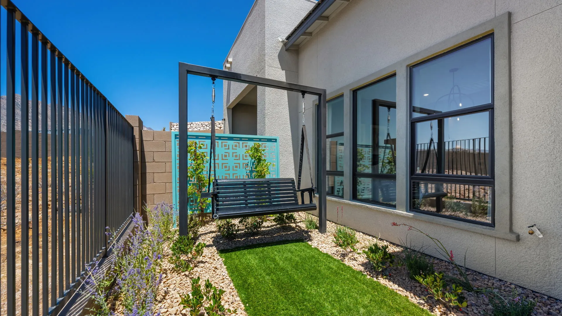 Backyard of Tiki Model at Esplanade at Red Rock by Taylor Morrison Black metal bench swing on green artificial grass beside a modern house, bordered by rocks and small plants. Tall fence and decorative screen provide privacy under a clear blue sky.