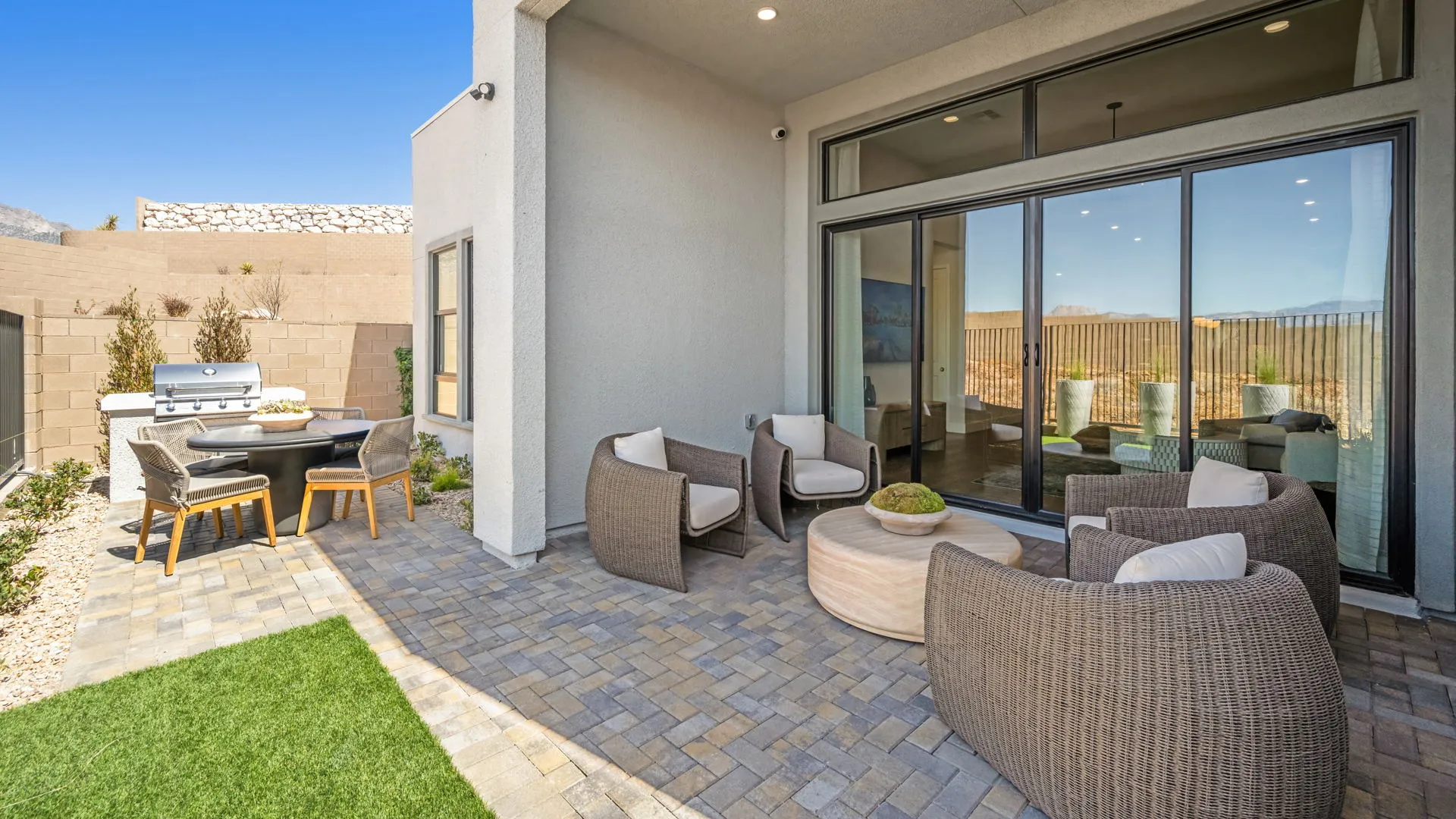 Backyard of Zinnia Model at Esplanade at Red Rock by Taylor Morrison (1) Modern backyard patio with wicker chairs, round tables, and a grill on paved flooring. Large glass doors open into the house; a lawn and stone wall surround the area under a clear blue sky.