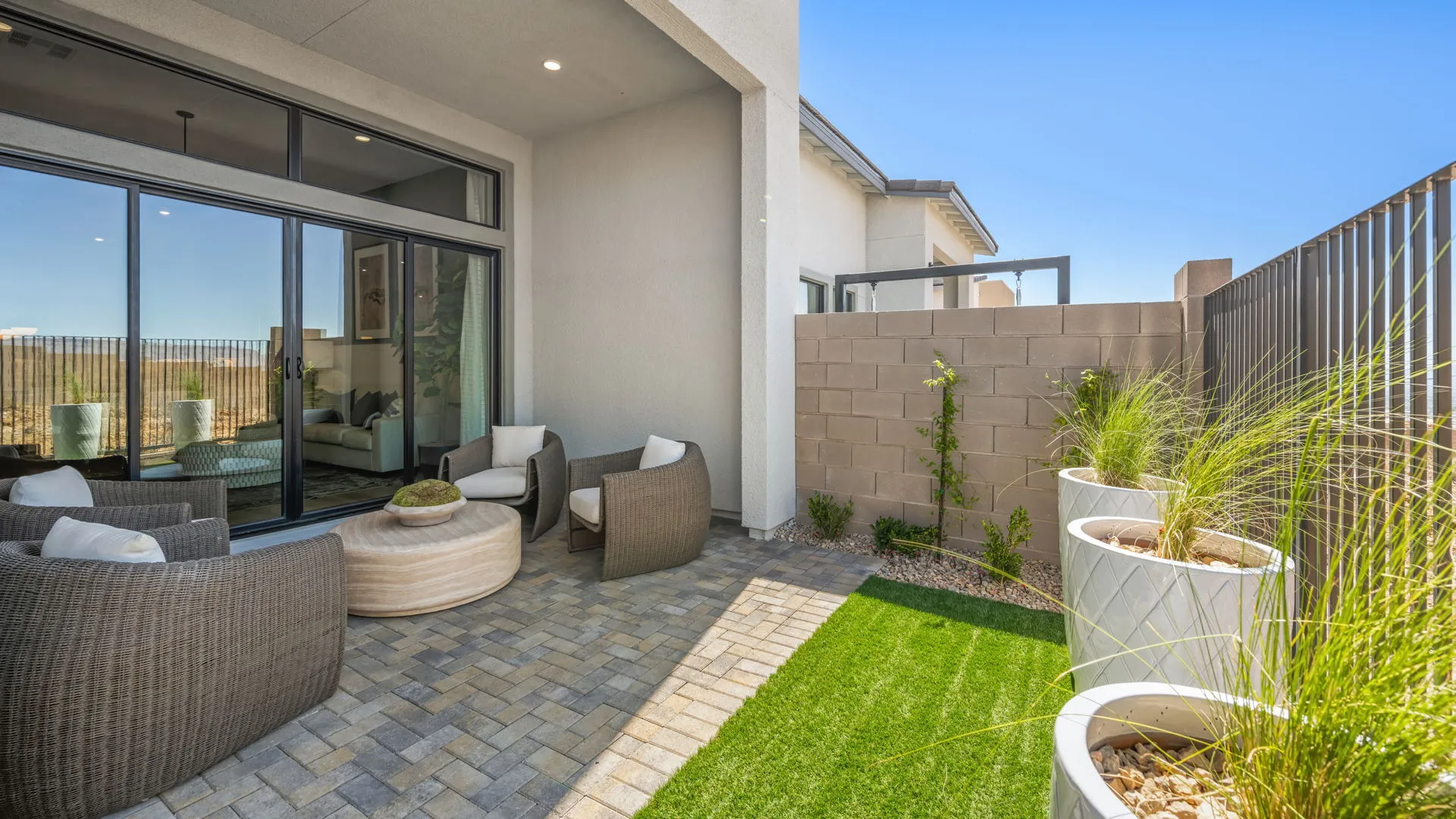 Backyard of Zinnia Model at Esplanade at Red Rock by Taylor Morrison (2) Modern patio with wicker chairs, a round ottoman, and potted plants on a paver floor. Sliding glass doors lead inside, and a stone wall separates the space from neighboring areas. Artificial grass adds greenery.