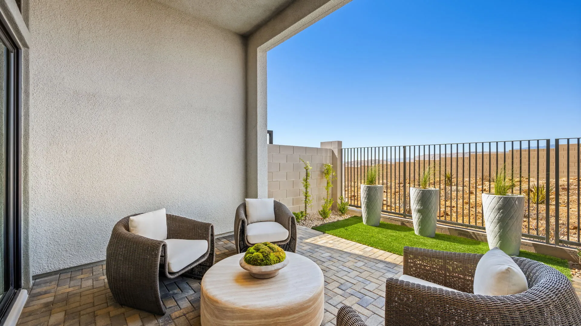 Backyard of Zinnia Model at Esplanade at Red Rock by Taylor Morrison (3) Covered patio with wicker chairs, a round table, and potted plants overlooks a small yard with artificial grass, a block wall, and metal fencing against a clear blue sky.
