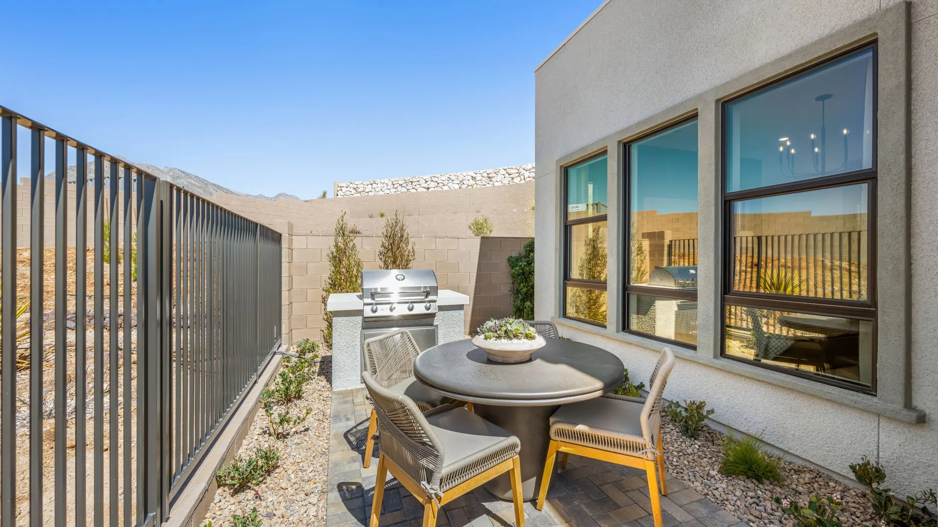 Backyard of Zinnia Model at Esplanade at Red Rock by Taylor Morrison A modern patio features a round table with four chairs, a built-in grill, potted plants, and a metal fence. Large windows reflect the clear blue sky.