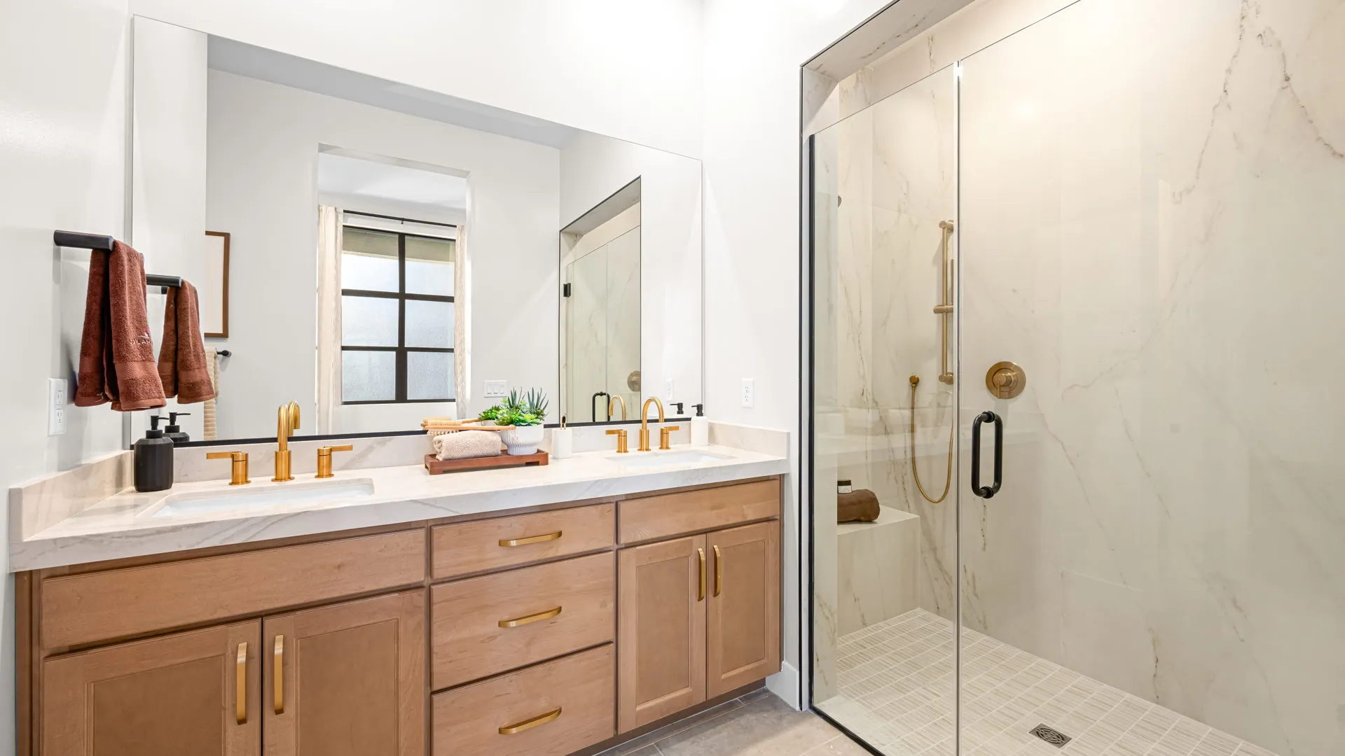 Bathroom of Monroe Model at Esplanade at Red Rock by Taylor Morrison (2) Modern bathroom with a glass shower, dual sinks with gold fixtures, wooden cabinets, white countertops, a large mirror, and folded towels on the counter. Neutral tones create a clean, bright look.