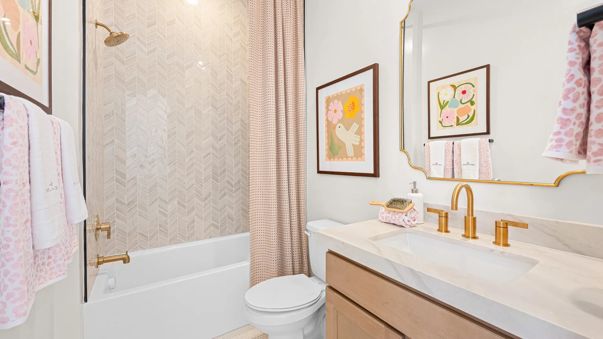 Bathroom of Monroe Model at Esplanade at Red Rock by Taylor Morrison Modern bathroom with a bathtub and shower, gold fixtures, light wood vanity, white countertop, pink patterned shower curtain, pastel floral art, and folded towels hanging on the wall and counter.