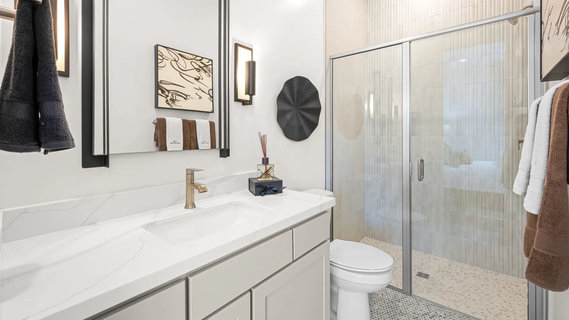 Bathroom of Paloma Model at Esplanade at Red Rock by Taylor Morrison Modern bathroom with white vanity, brass faucet, wall art, towels, and glass-enclosed shower. Neutral tones and minimalist decor create a clean, elegant look.
