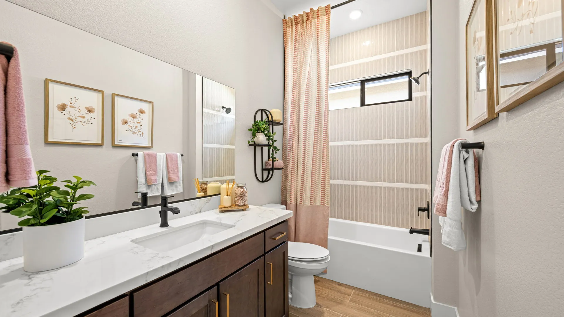 Bathroom of Sangria Model at Esplanade at Red Rock by Taylor Morrison (1) Modern bathroom with a marble countertop, dark wood cabinets, wall art, a white sink, pink towels, a potted plant, shelving, and a bathtub with a pink curtain and window. Light walls and wood-look floor.