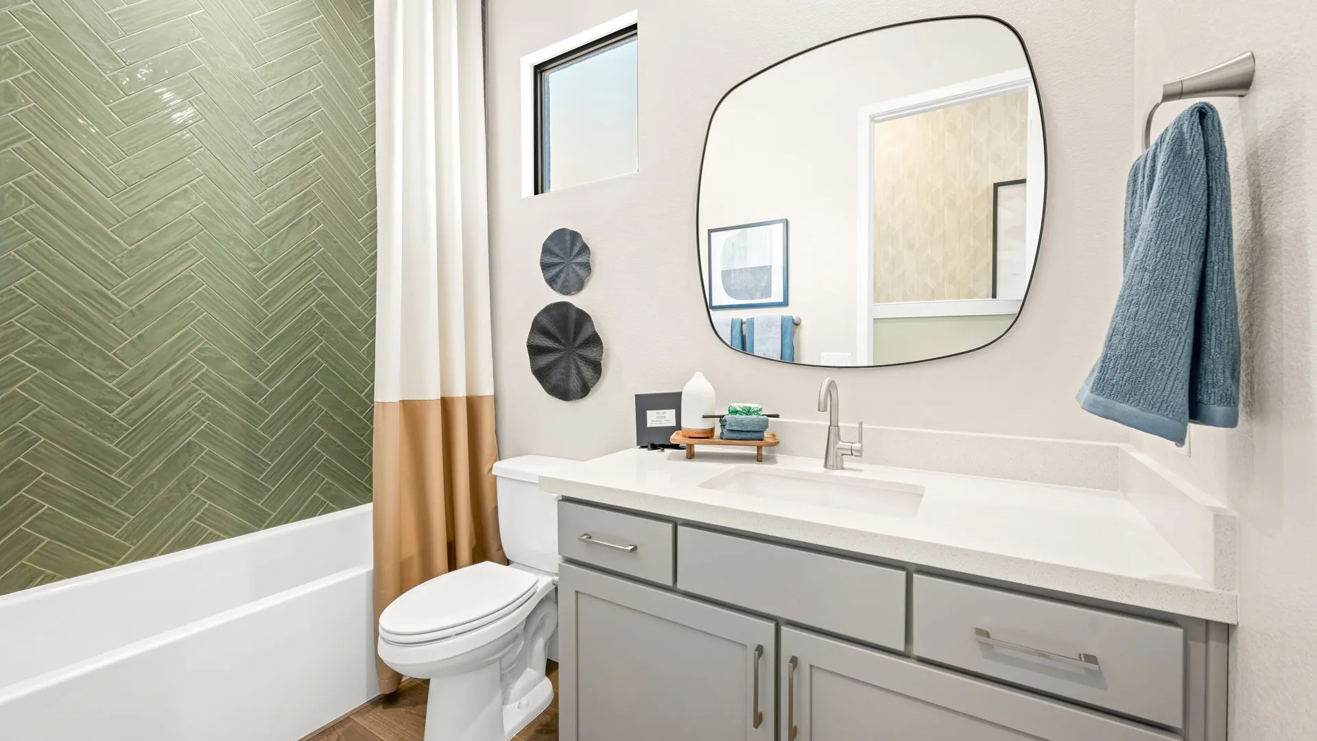 Bathroom of Zinnia Model at Esplanade at Red Rock by Taylor Morrison Modern bathroom with green herringbone tile in the shower, a white and tan shower curtain, gray vanity, oval mirror, white countertop, and a blue towel hanging on the wall.