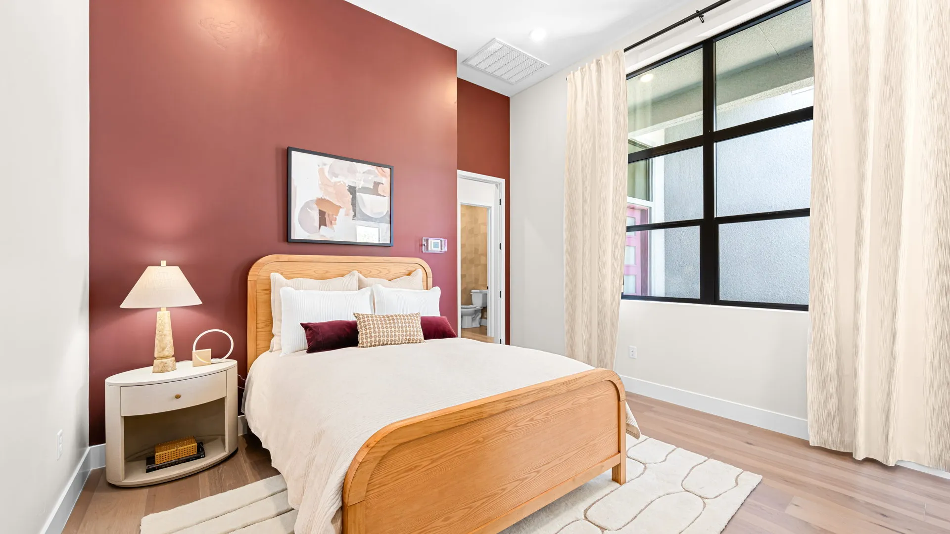 Bedroom of Monroe Model at Esplanade at Red Rock by Taylor Morrison (2) A cozy bedroom with a wooden bed, cream bedding, maroon accent wall, abstract art, nightstand with lamp, large window with beige curtains, and light wood flooring.