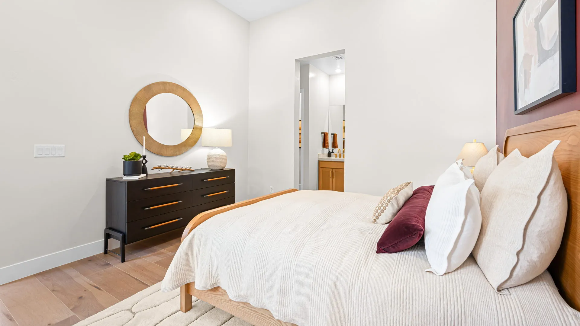 Bedroom of Monroe Model at Esplanade at Red Rock by Taylor Morrison (37) A modern bedroom features a wooden bed with cream bedding, accent pillows, a dark dresser with a round mirror, a lamp, and a plant. Light walls and wood flooring create a bright, cozy atmosphere.
