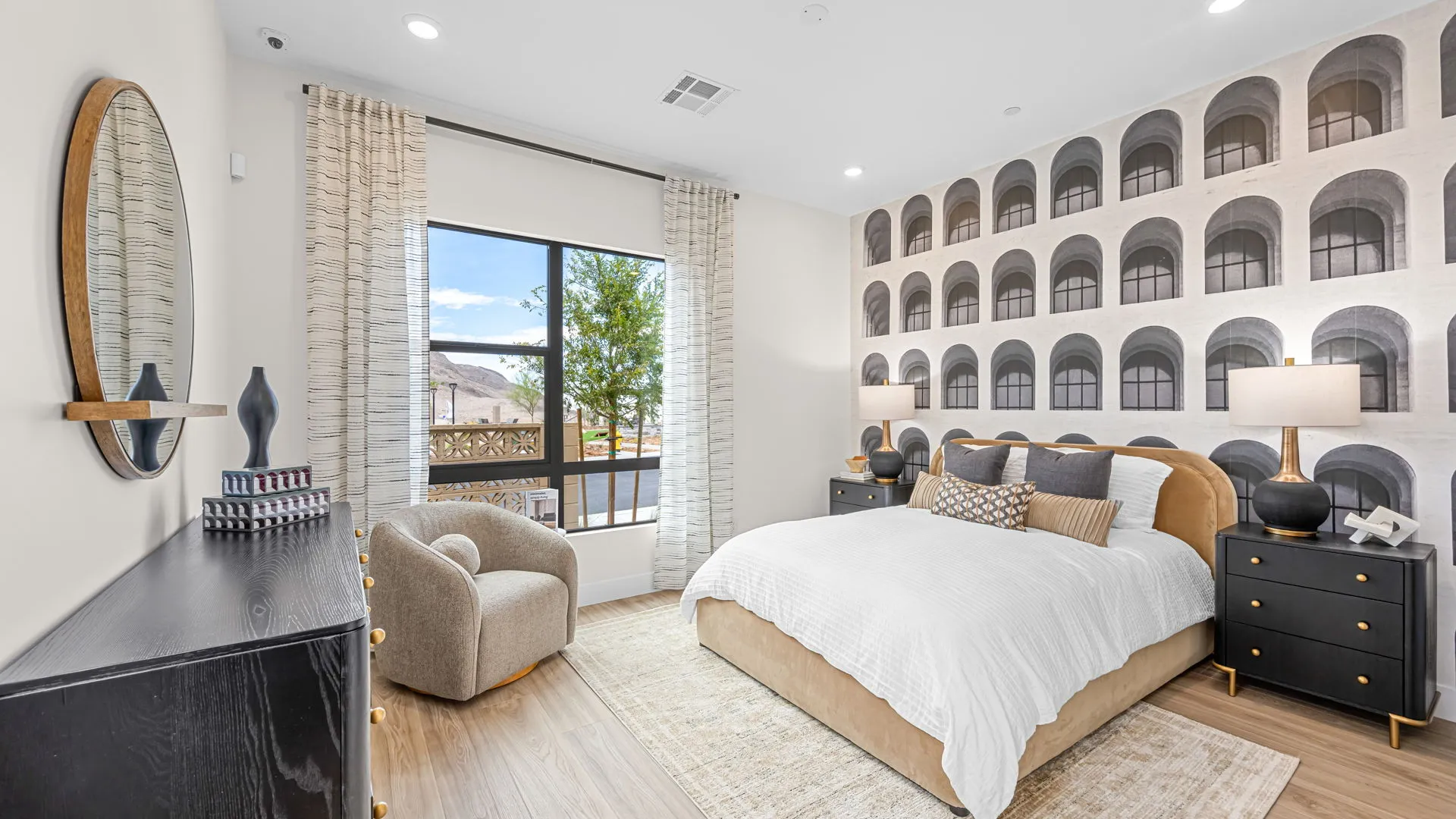 Bedroom of Paloma Model at Esplanade at Red Rock by Taylor Morrison (1) Modern bedroom with a large window, bed with white bedding, unique wall mural of arches, black nightstands, a round mirror, and a cozy armchair in the corner.