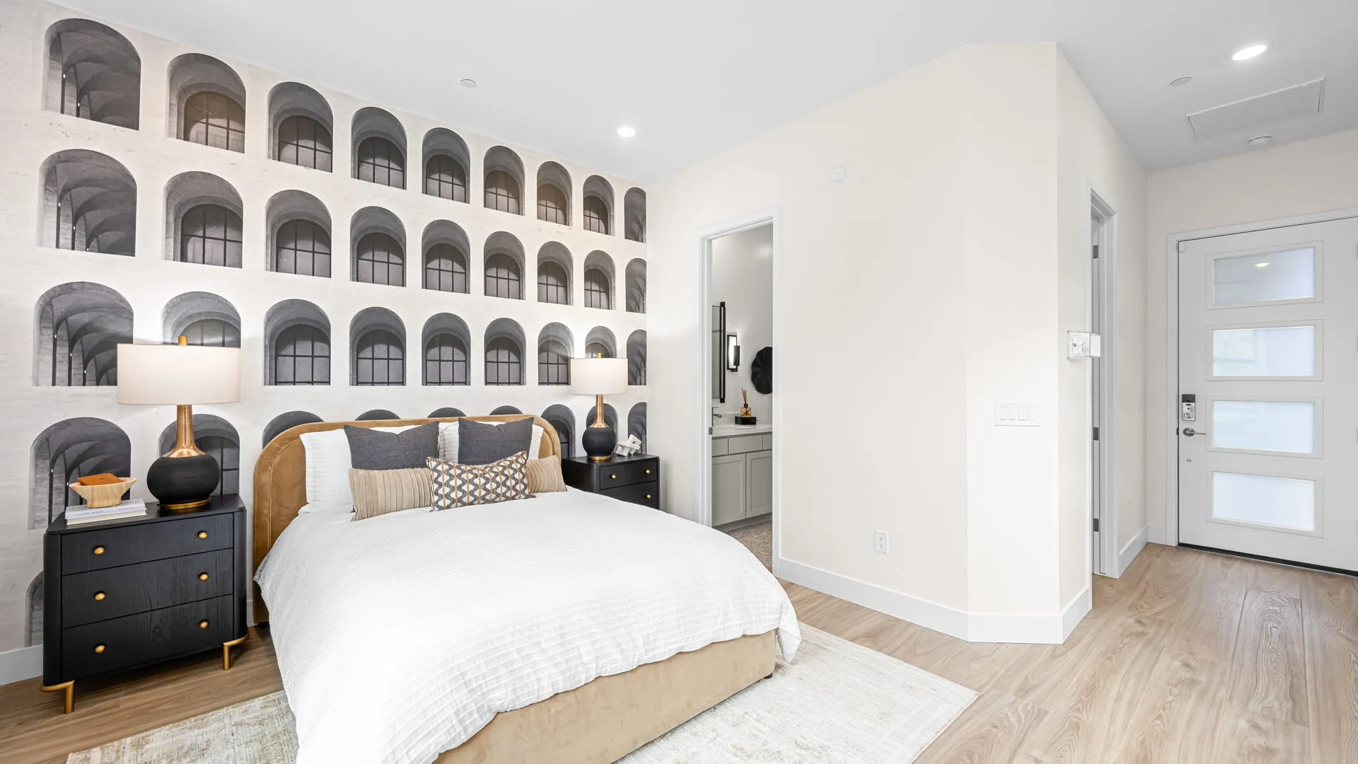 Bedroom of Paloma Model at Esplanade at Red Rock by Taylor Morrison Modern bedroom with a patterned accent wall featuring arches, a bed with white bedding, black nightstands with lamps, and a doorway leading to a bathroom. Light wood flooring and minimalist decor.