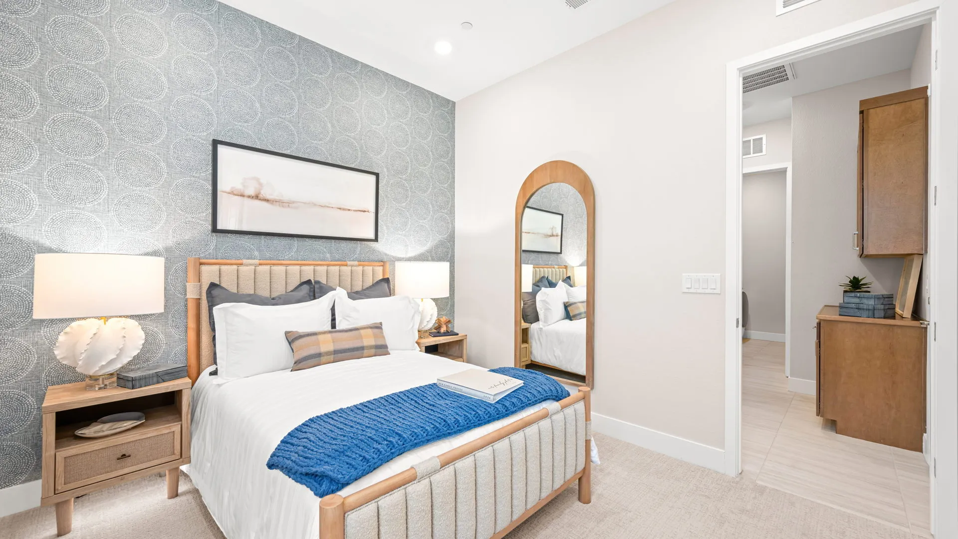 Bedroom of Tiki Model at Esplanade at Red Rock by Taylor Morrison (1) Modern bedroom with a wooden bed, white bedding, blue throw, two nightstands with lamps, a large mirror, and a patterned accent wall. An open doorway leads to another room with cabinetry.