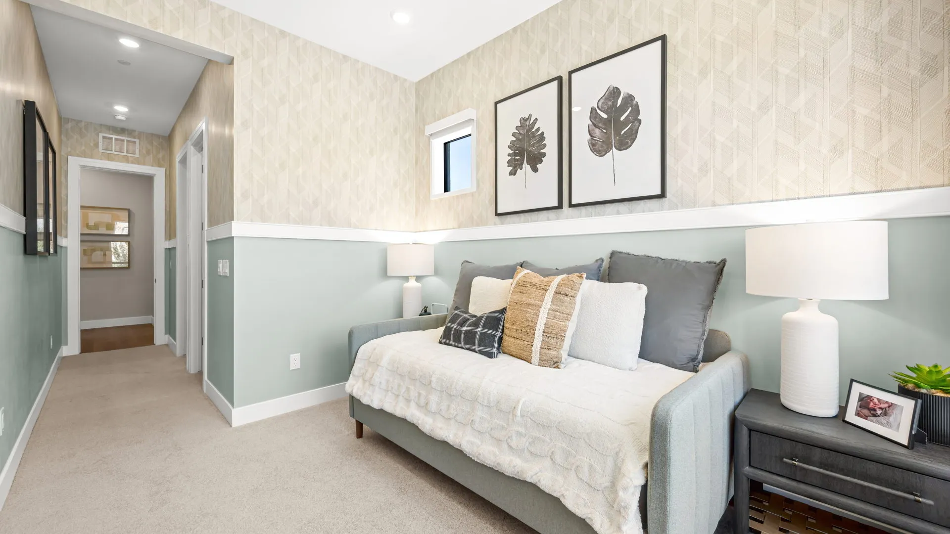 Bedroom of Zinnia Model at Esplanade at Red Rock by Taylor Morrison (1) A cozy, light-filled room with a daybed, decorative pillows, two lamps, framed leaf art, a nightstand, and patterned wallpaper; a hallway and doors are visible in the background.