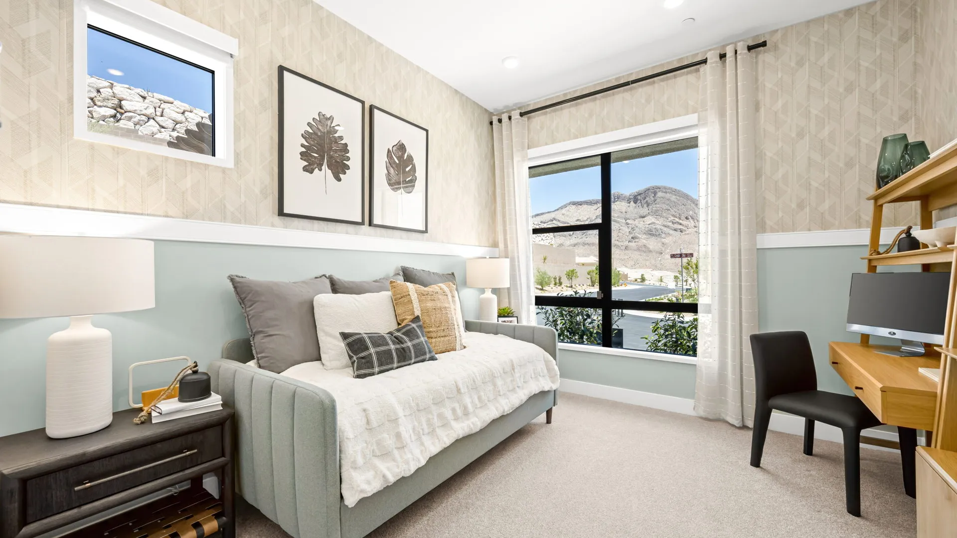 Bedroom of Zinnia Model at Esplanade at Red Rock by Taylor Morrison Bright bedroom with a daybed, throw pillows, side table, two lamps, wall art, desk with a computer, black chair, large window with mountain view, and neutral decor.