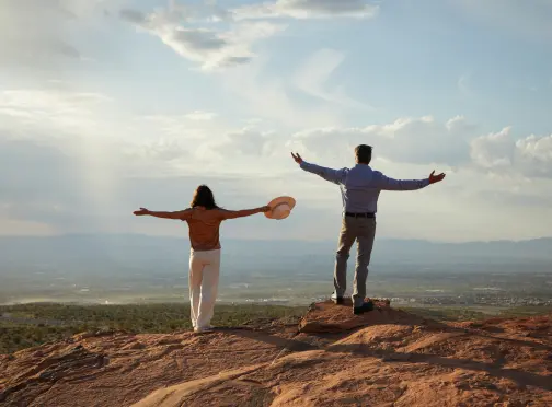 A man and woman stand on a rocky hilltop with arms outstretched, facing a scenic view of open landscape and distant mountains under a partly cloudy sky.