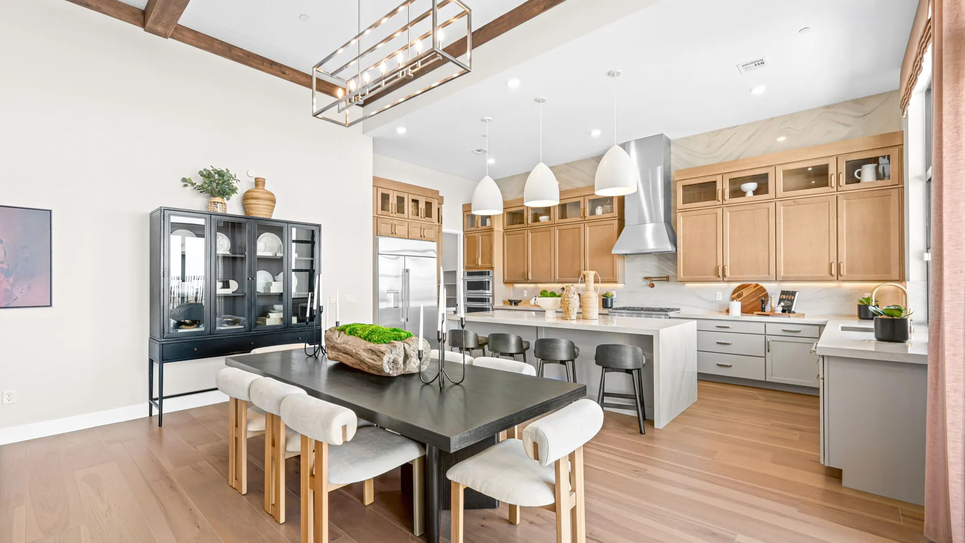Dining Room of Monroe Model at Esplanade at Red Rock by Taylor Morrison (1) Modern kitchen and dining area with light wood cabinets, stainless steel appliances, a black dining table with six chairs, black display cabinet, pendant lights, and decorative plants on wood flooring.