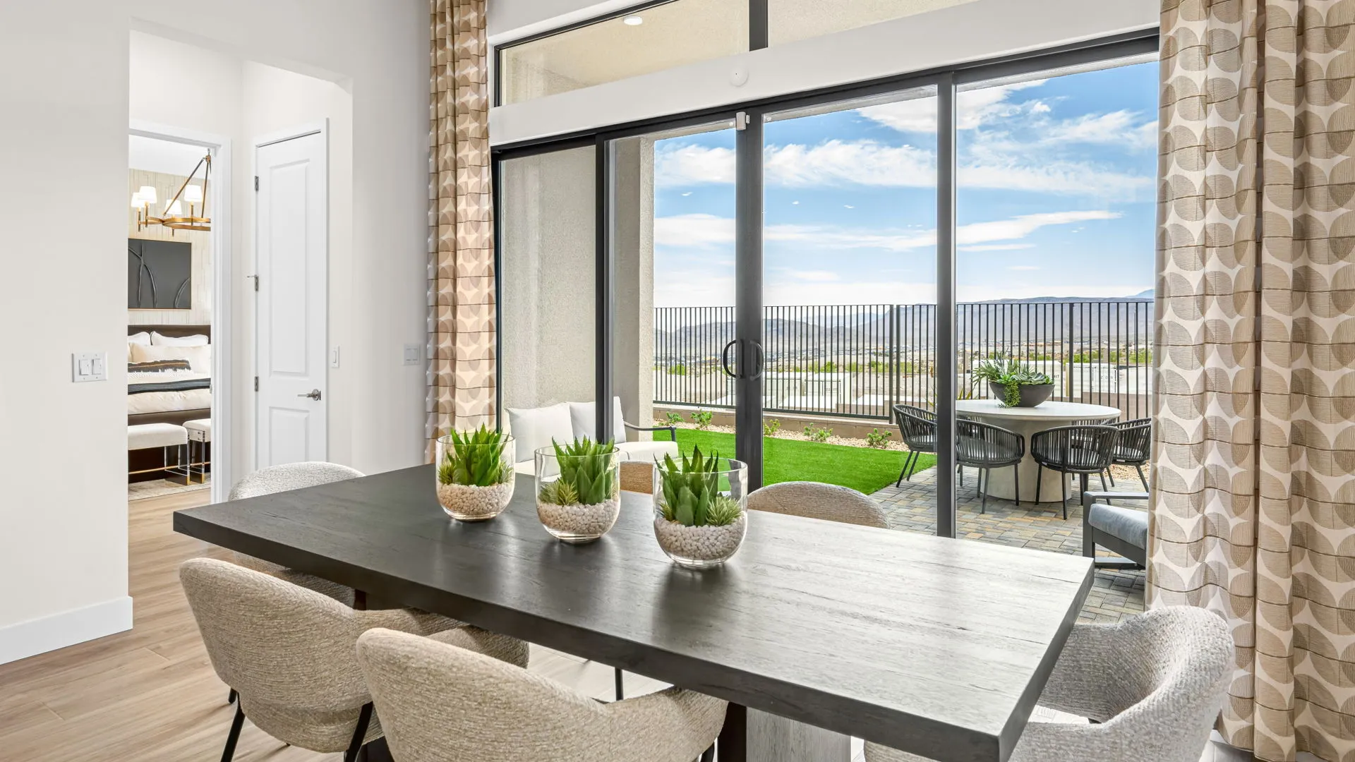 Dining Room of Paloma Model at Esplanade at Red Rock by Taylor Morrison Modern dining area with a dark table, light chairs, and potted plants, next to large glass doors that open to a patio with outdoor seating and a green lawn, overlooking a scenic, fenced landscape under a blue sky.