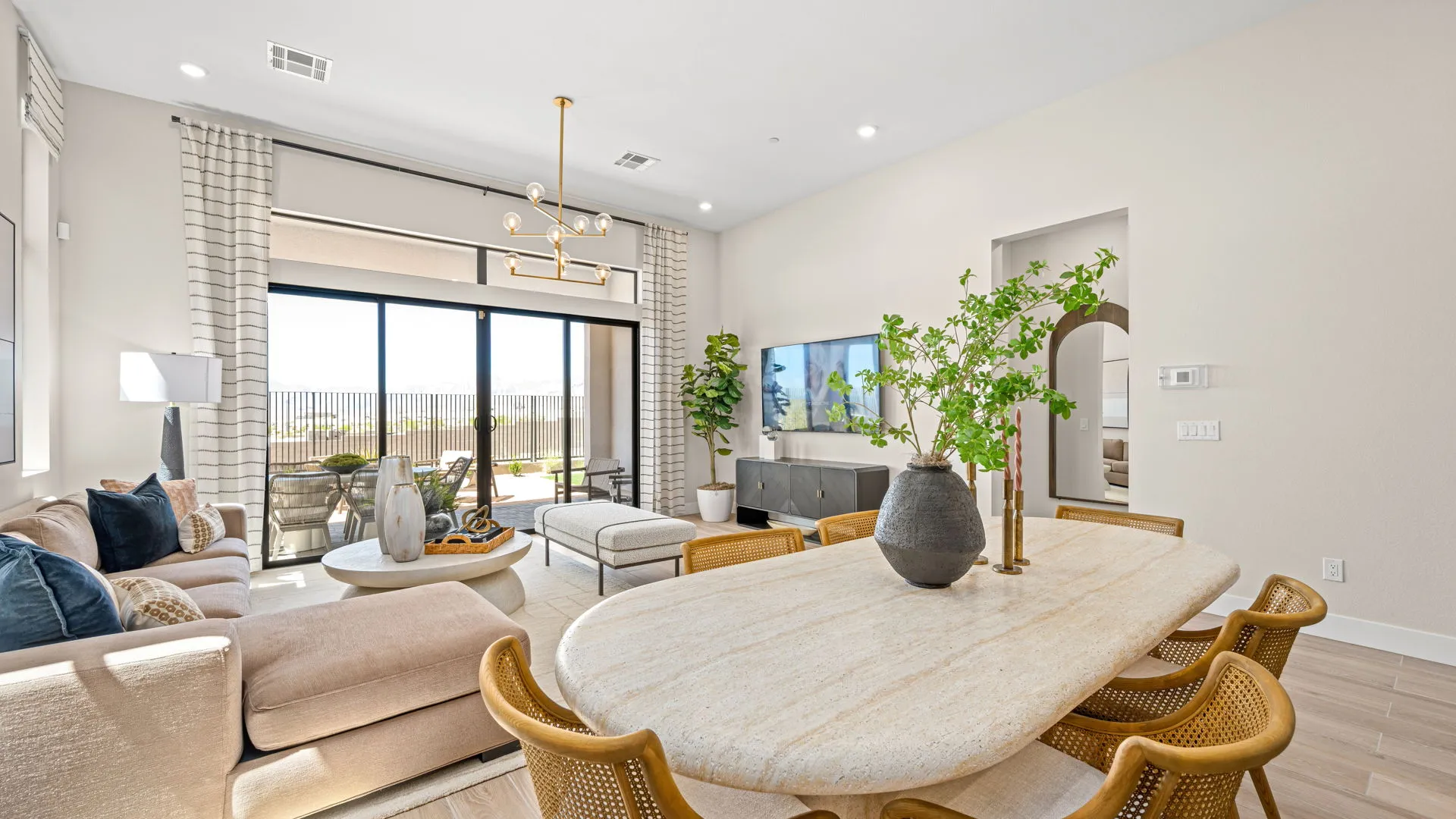 Dining Room of Sangria Model at Esplanade at Red Rock by Taylor Morrison Modern living and dining area with beige walls, light furniture, a marble dining table, green plants, a large TV, and sliding glass doors leading to a patio with outdoor seating and a scenic view.