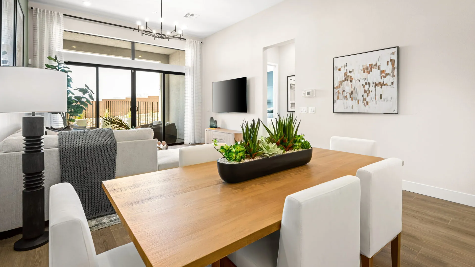 Dining Room of Zinnia Model at Esplanade at Red Rock by Taylor Morrison (1) Modern dining and living area with a wooden table, white chairs, sofa, TV on the wall, large windows letting in natural light, and decorative plants as a centerpiece.