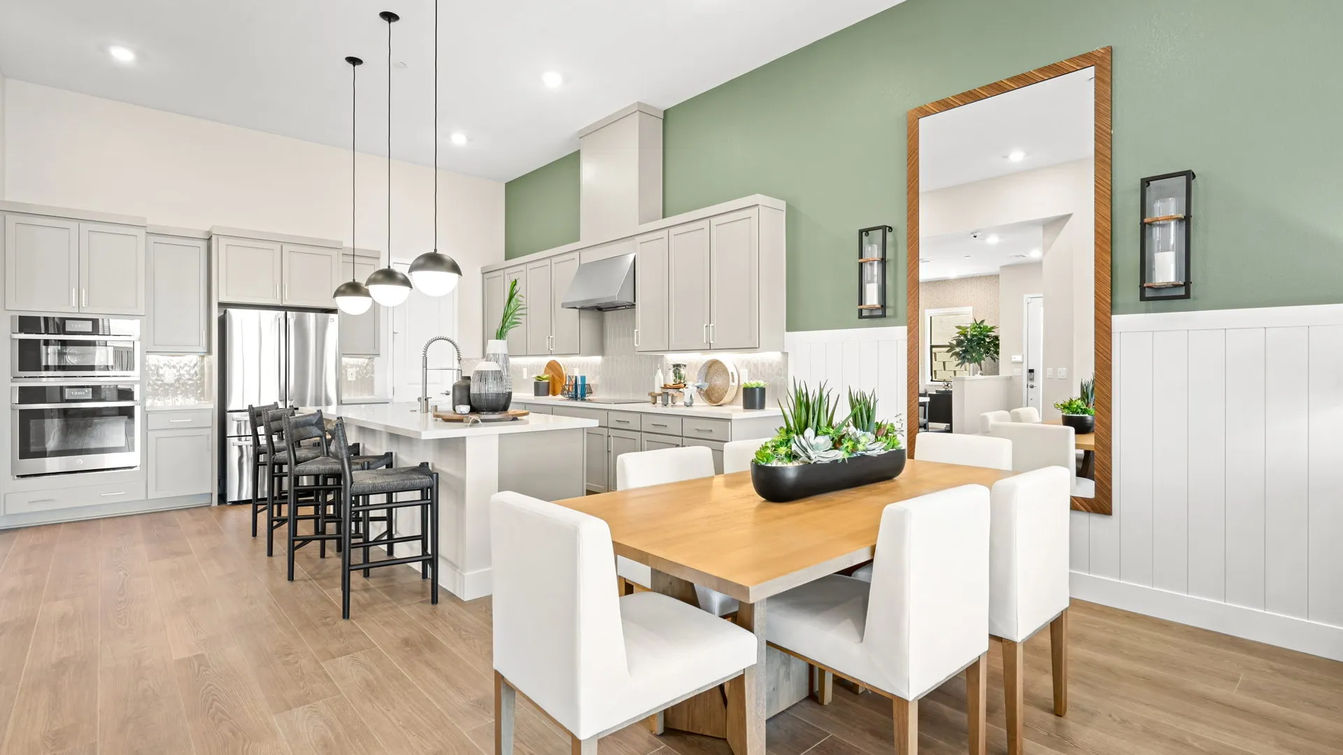 Dining Room of Zinnia Model at Esplanade at Red Rock by Taylor Morrison (3) Modern kitchen and dining area with light wood flooring, green accent wall, white cabinetry, a large mirror, pendant lights, and a wooden dining table surrounded by white chairs with a plant centerpiece.