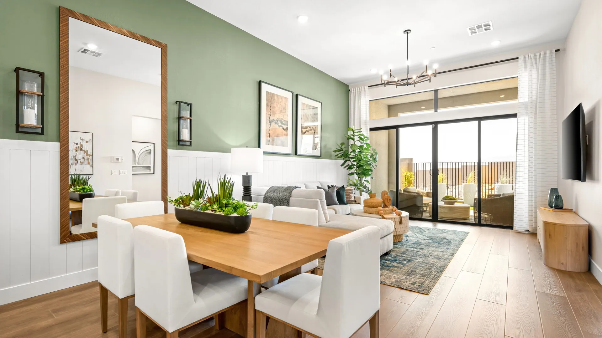 Dining Room of Zinnia Model at Esplanade at Red Rock by Taylor Morrison Modern open living-dining area with green accent wall, wood dining table, white chairs, beige sofa, large window, indoor plants, wall art, and natural light.