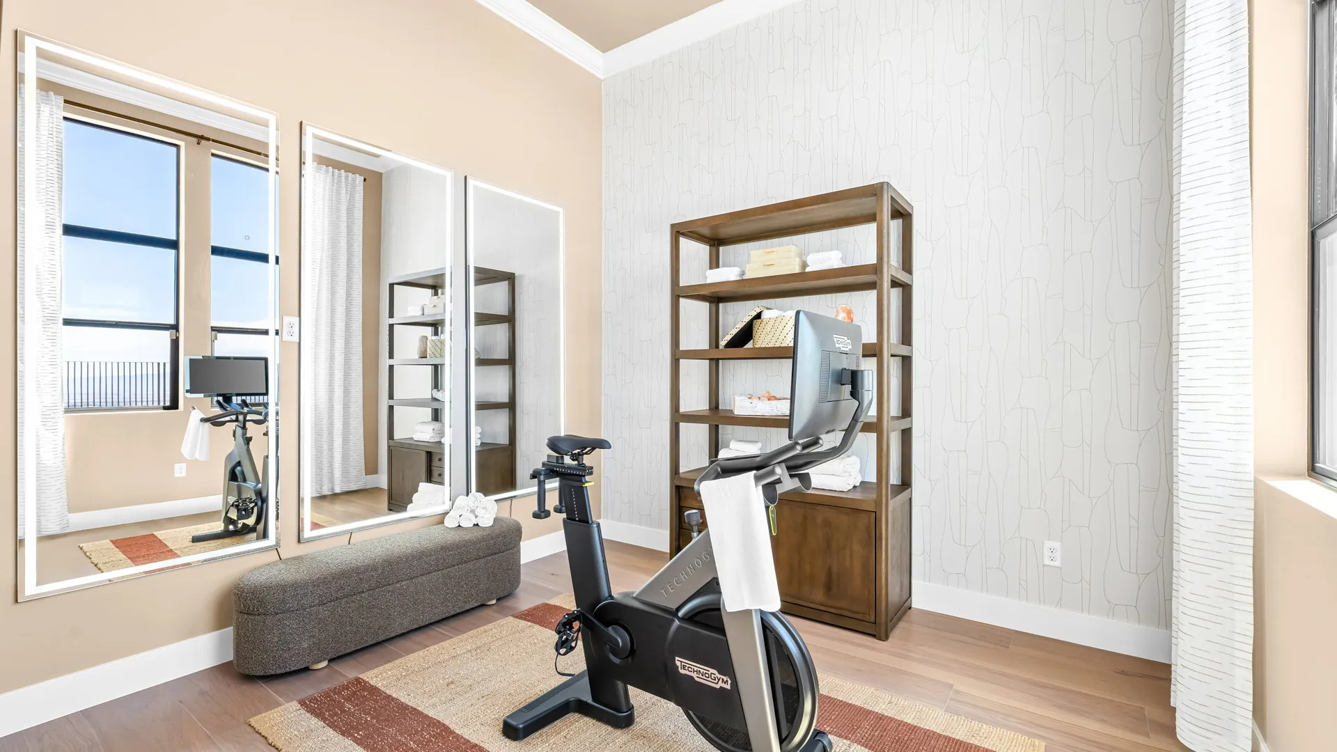 Exercise Room of Monroe Model at Esplanade at Red Rock by Taylor Morrison A bright home gym features an exercise bike on a rug, a brown bench near three tall mirrors, two wooden shelves with towels and decor, and large windows letting in natural light.