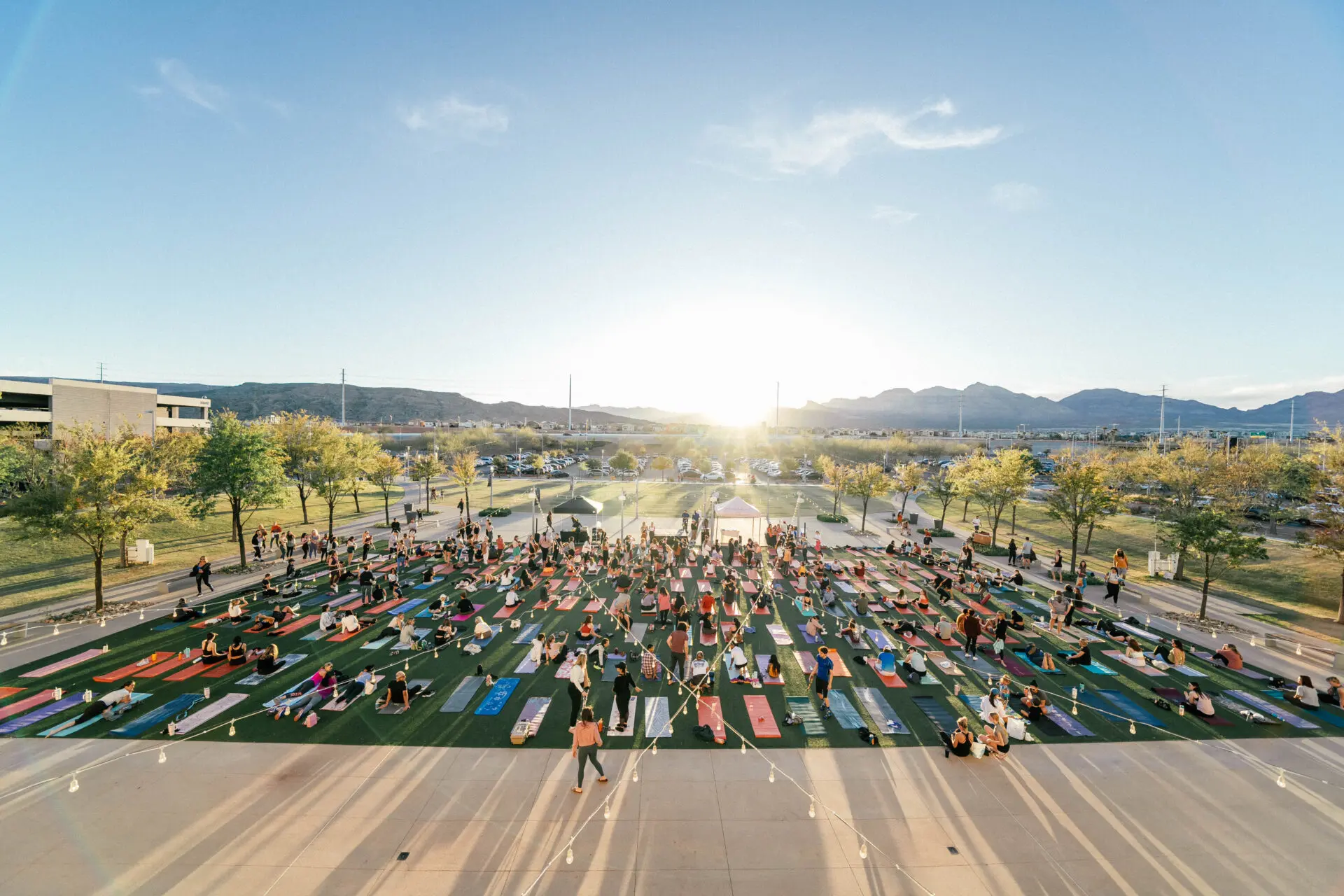 A large outdoor yoga class takes place at sunset, with many people on colorful mats spread across a green field, surrounded by trees and distant mountains.