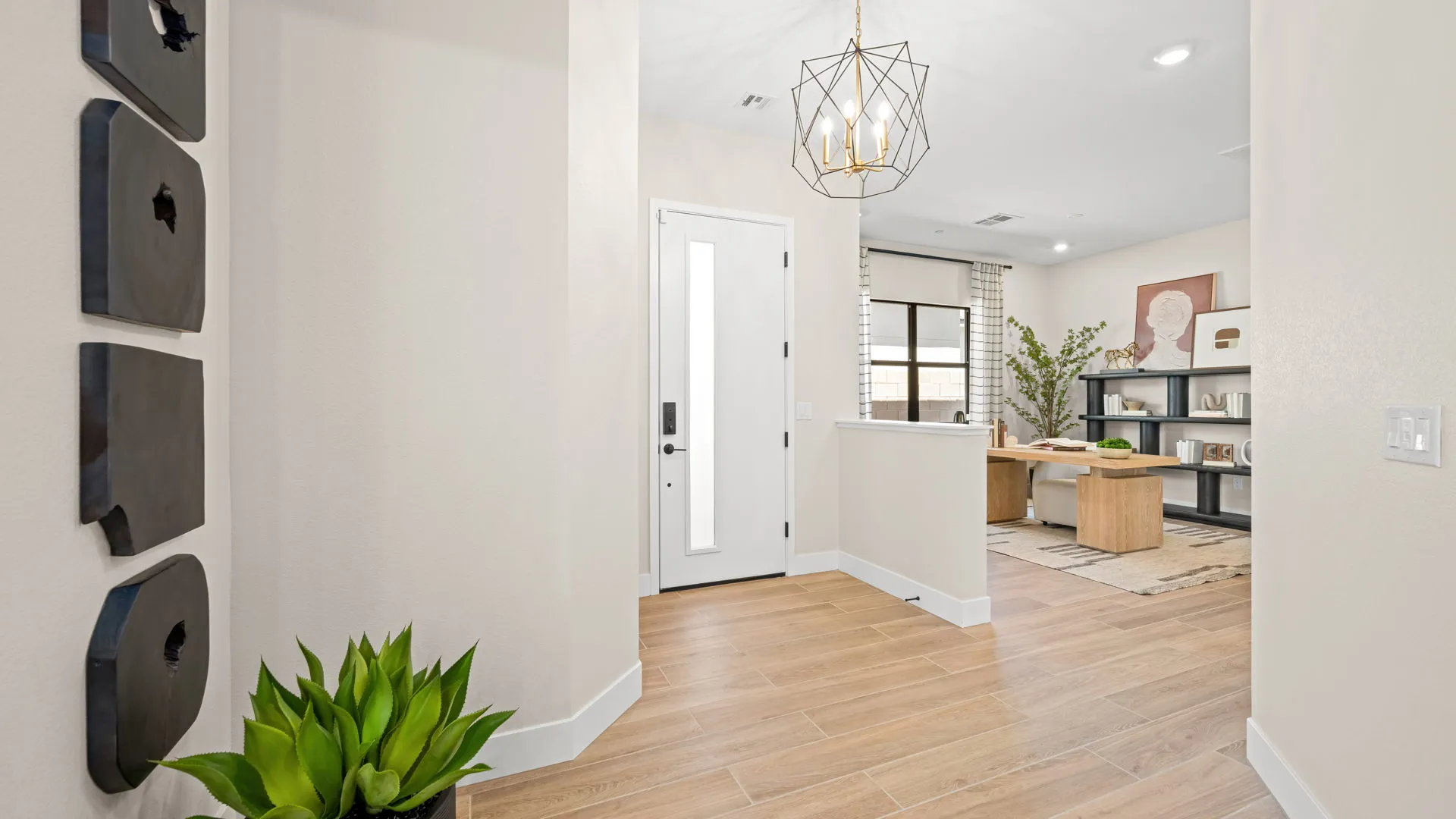 Foyer of Sangria Model at Esplanade at Red Rock by Taylor Morrison A bright hallway with light wood floors, a potted plant, wall art, and a modern chandelier leads to a home office with shelves, a desk, and a large window.