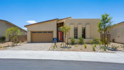 Modern single-story house with a tan exterior, two-car garage, red front door, and xeriscaped yard featuring small trees and rocks under a clear blue sky.
