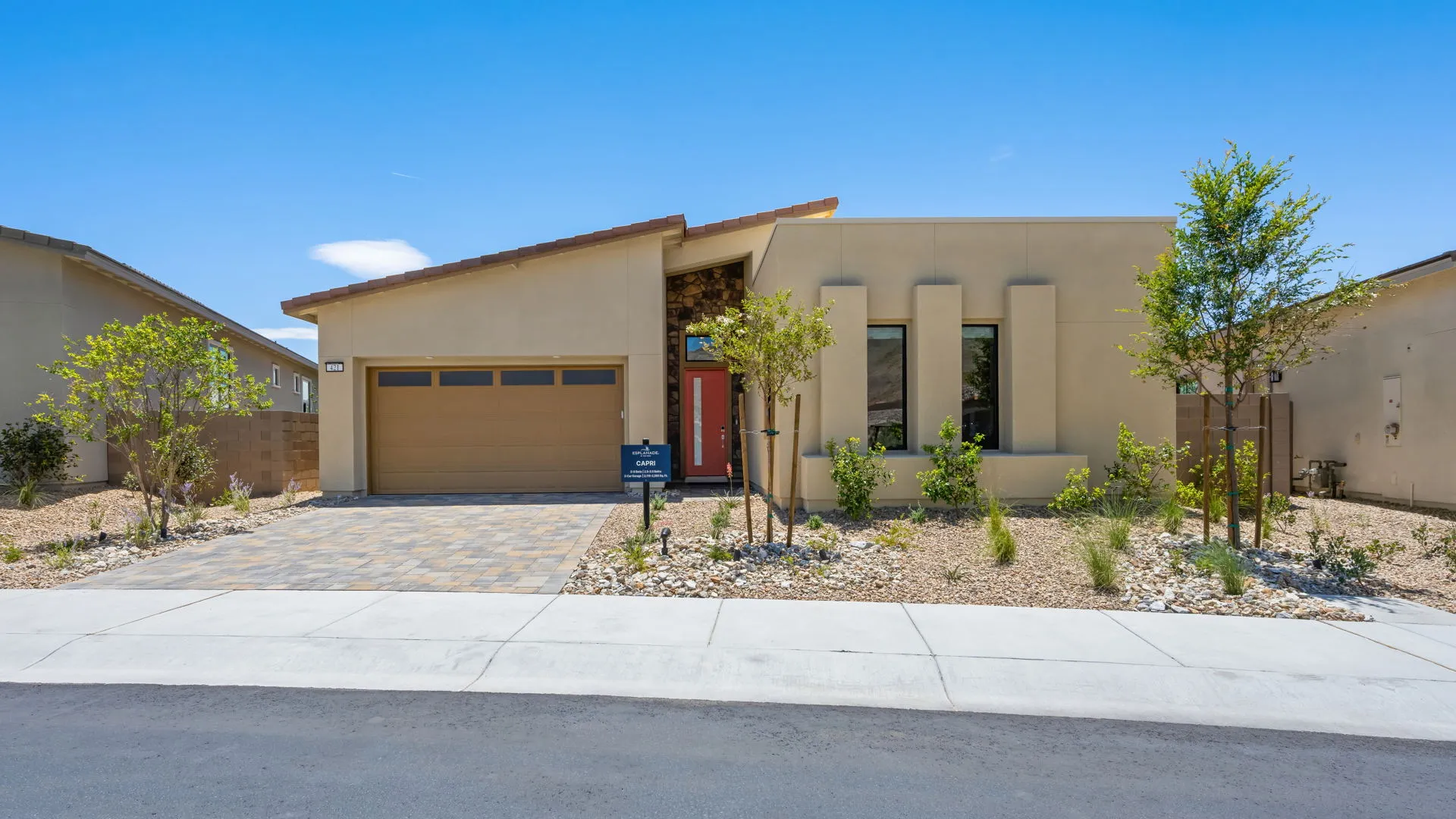 Modern single-story house with a tan exterior, two-car garage, red front door, and xeriscaped yard featuring small trees and rocks under a clear blue sky.