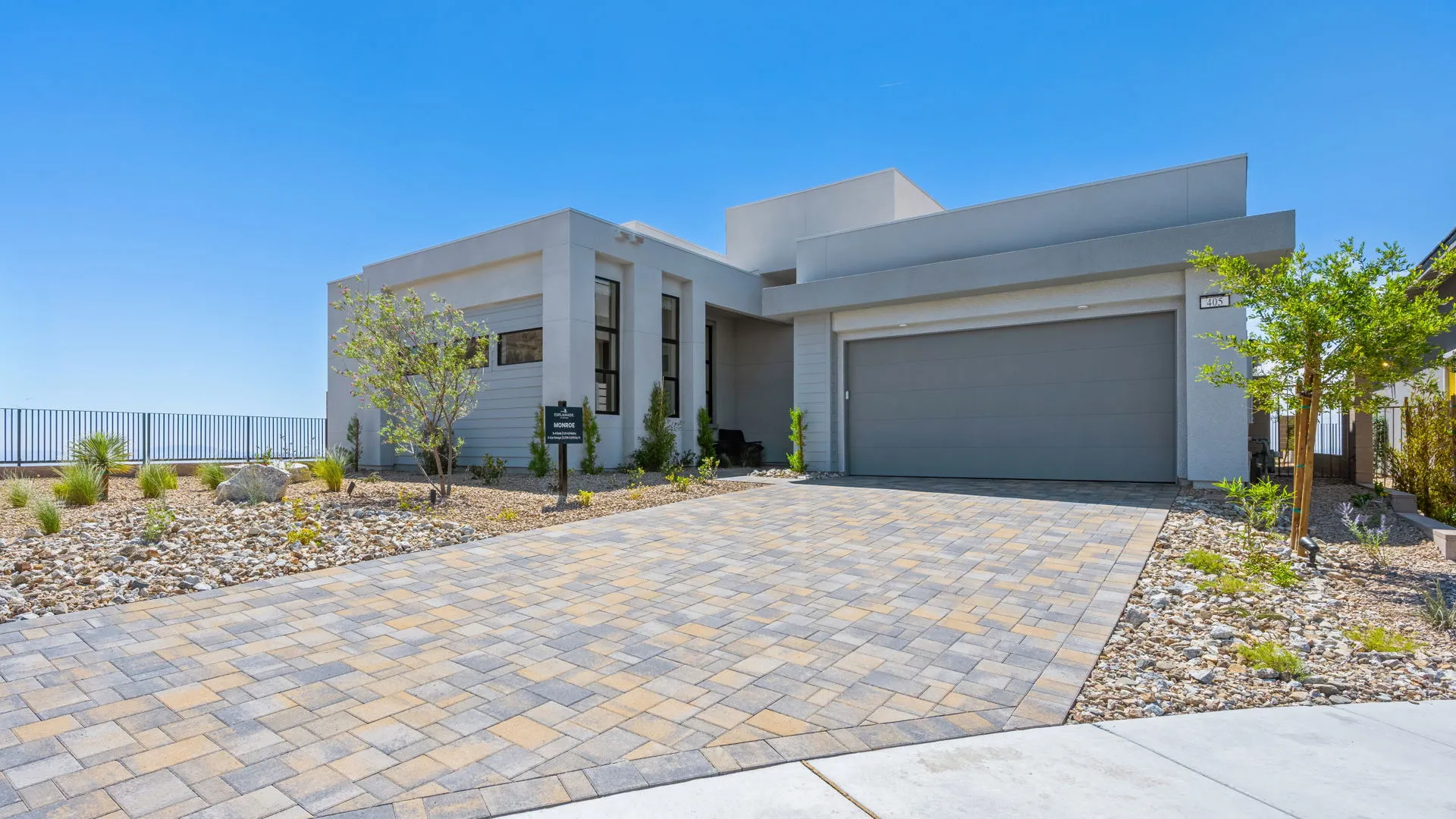 Front Elevation of Monroe Model at Esplanade at Red Rock by Taylor Morrison (1) A modern, single-story house with a flat roof, light gray exterior, two-car garage, and a wide paved driveway surrounded by desert landscaping under a clear blue sky.