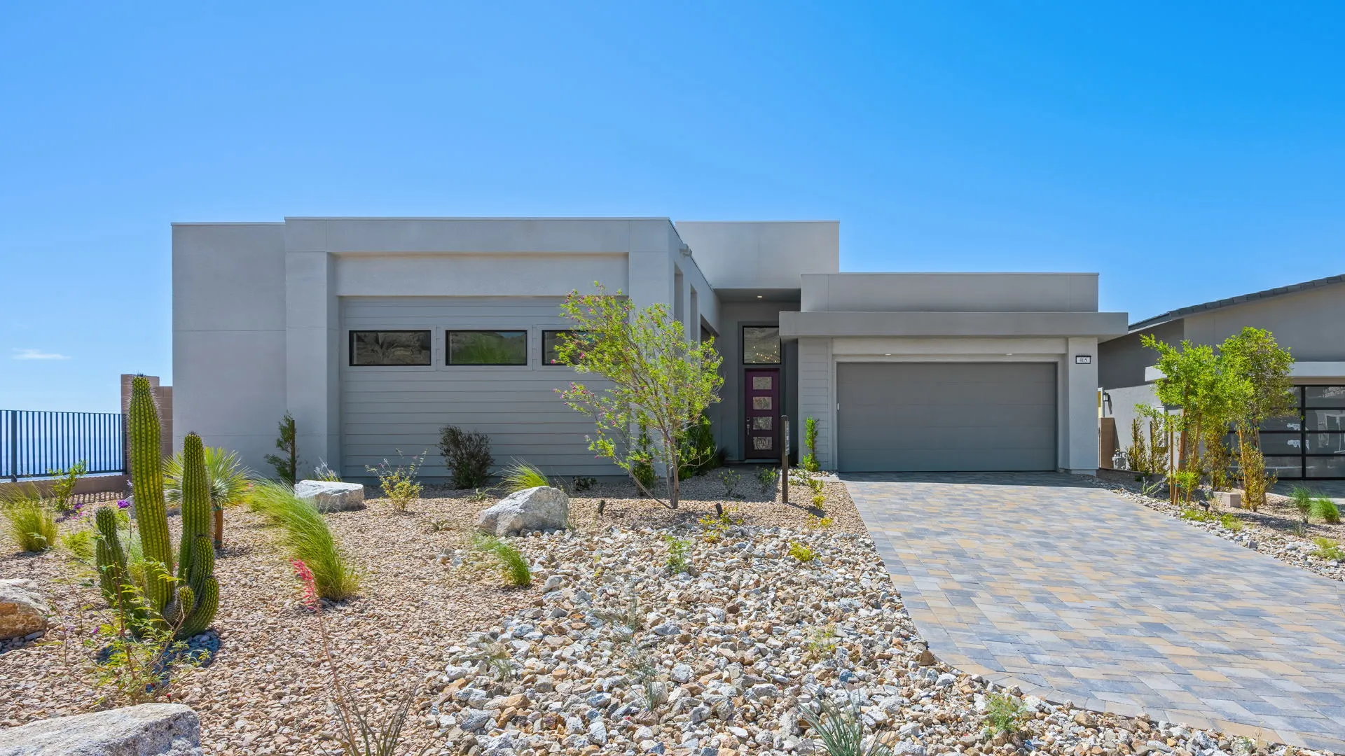 Front Elevation of Monroe Model at Esplanade at Red Rock by Taylor Morrison Modern single-story house with flat roof, light gray exterior, two-car garage, and a purple front door. The front yard features desert landscaping with rocks, cacti, and small trees under a clear blue sky.