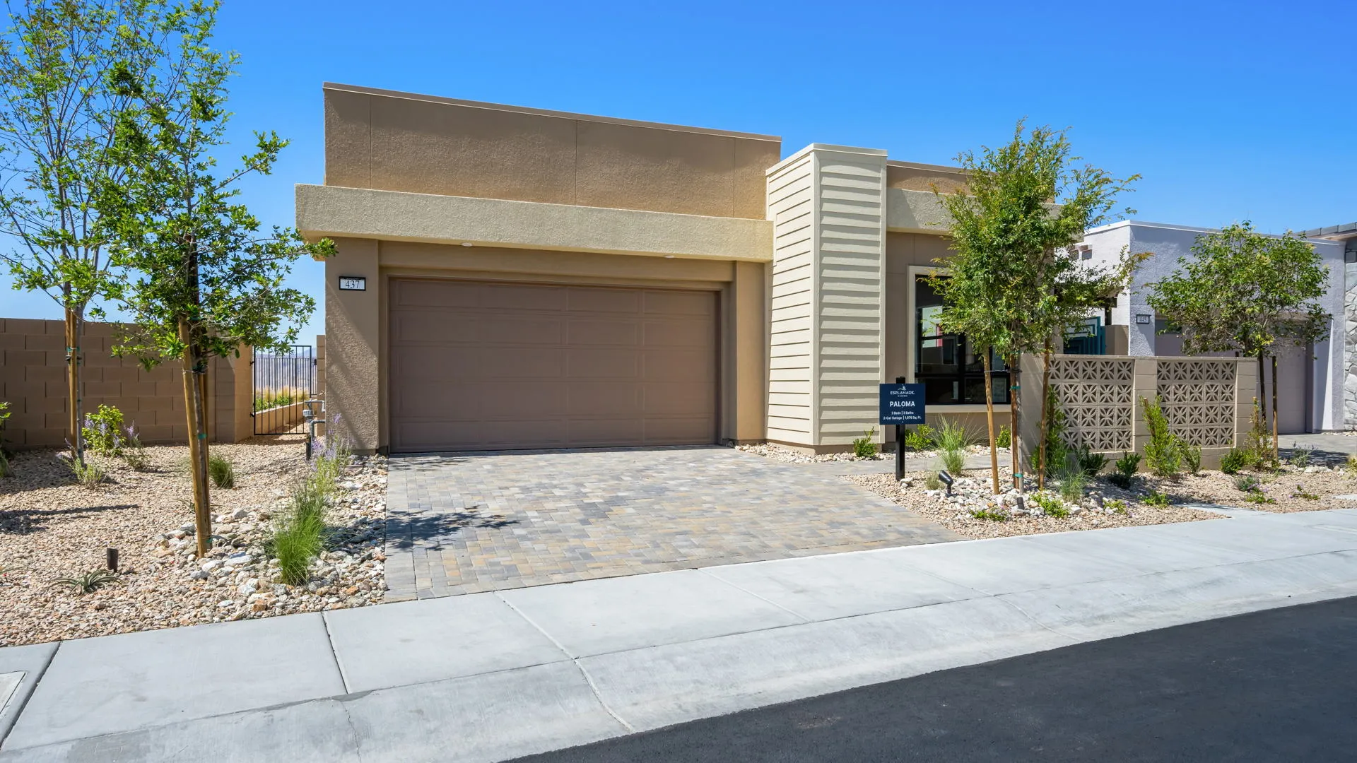 Front Elevation of Paloma Model at Esplanade at Red Rock by Taylor Morrison (1) Modern single-story house with a tan exterior, two-car garage, paved driveway, minimal desert landscaping, and small trees in front, set under a clear blue sky.
