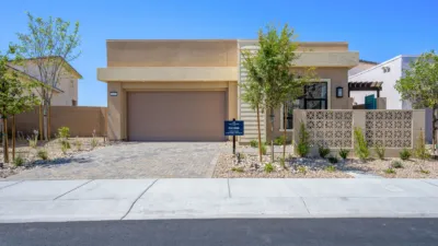 Single-story modern house with a flat roof, beige exterior, two-car garage, and minimal desert landscaping. A small tree and sign are near the walkway leading to the front entrance. Blue sky in the background.