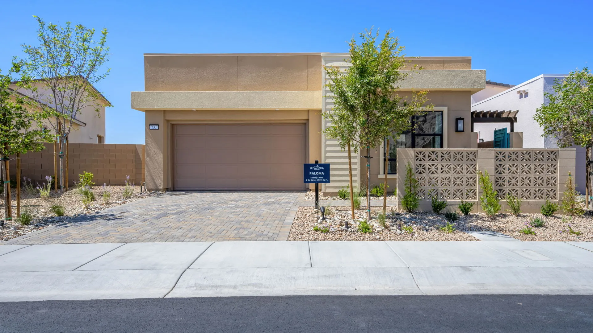 Front Elevation of Paloma Model at Esplanade at Red Rock by Taylor Morrison Single-story modern house with a flat roof, beige exterior, two-car garage, and minimal desert landscaping. A small tree and sign are near the walkway leading to the front entrance. Blue sky in the background.