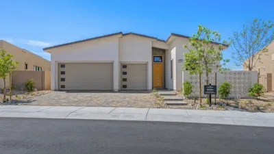 Single-story modern house with a flat roof, two-car garage, beige exterior, and a yellow front door. The yard features stone landscaping, small trees, and a model home sign near the sidewalk.