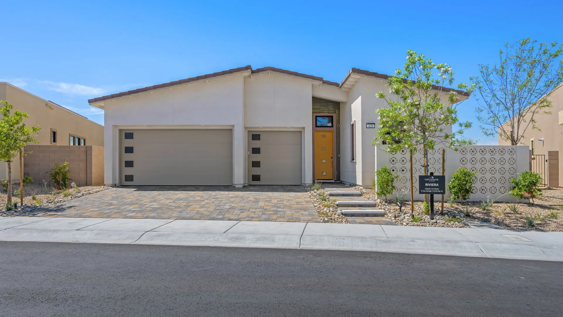 Single-story modern house with a flat roof, two-car garage, beige exterior, and a yellow front door. The yard features stone landscaping, small trees, and a model home sign near the sidewalk.
