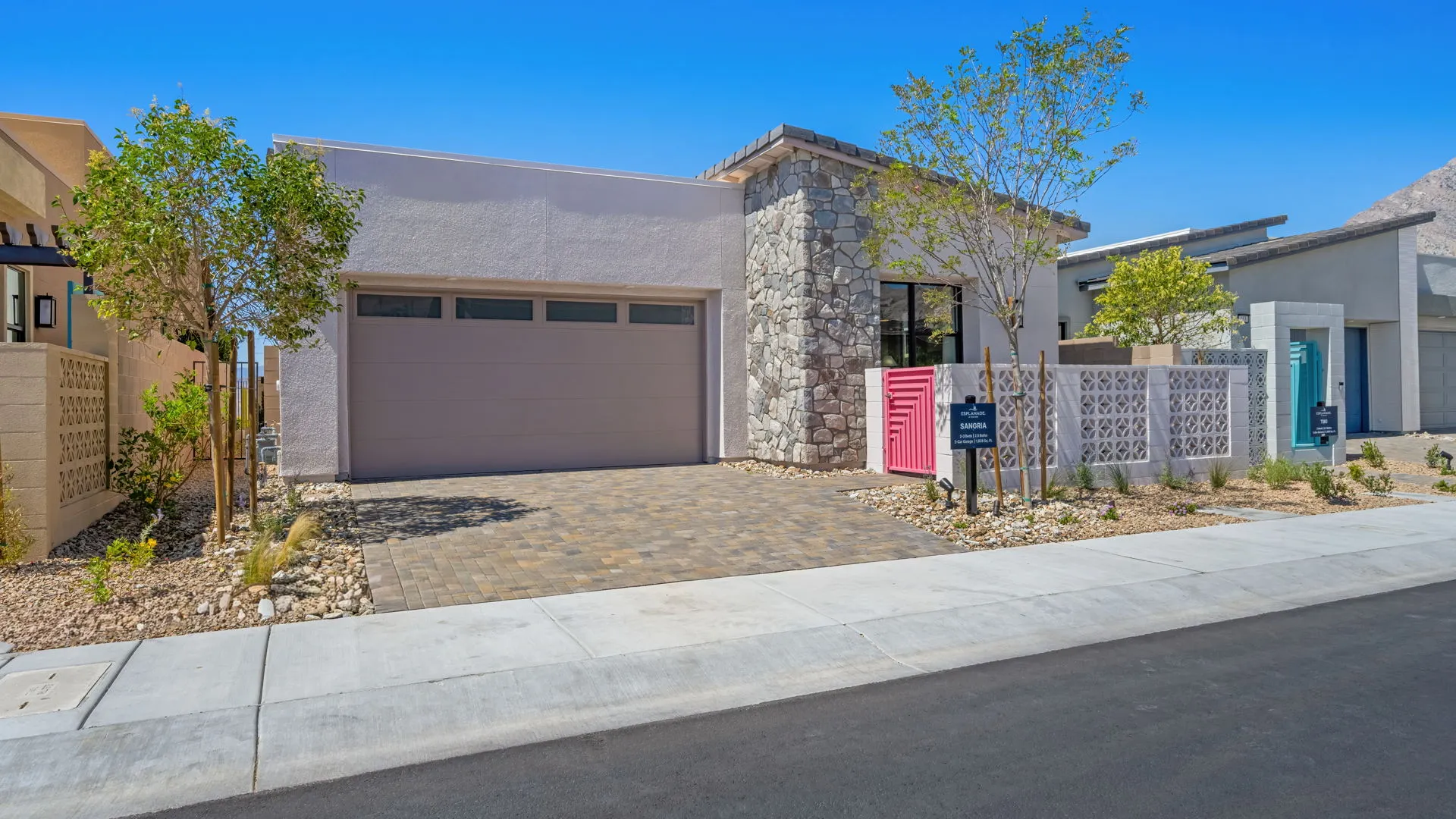 Front Elevation of Sangria Model at Esplanade at Red Rock by Taylor Morrison (1) Modern single-story house with a stone and stucco exterior, double garage, pink front door, desert landscaping, and young trees, set on a paved street under a clear blue sky.