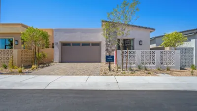 Modern single-story house with a flat roof, two-car garage, decorative concrete wall, small trees, and desert landscaping under a clear blue sky.